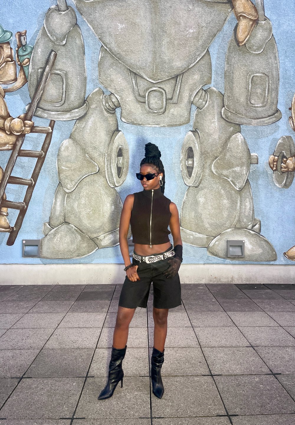 Top preto sem mangas com fecho, calções pretos de cintura alta e botas pretas até ao joelho; cabelo trançado; de pé à frente de um mural.
