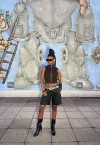 Top preto sem mangas com fecho, calções pretos de cintura alta e botas pretas até ao joelho; cabelo trançado; de pé à frente de um mural.