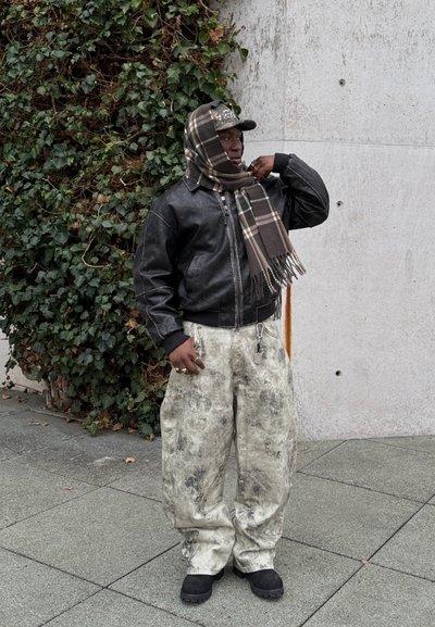 Chaqueta de cuero negra, bufanda marrón con estampado, pantalones ligeros grises grandes con manchas oscuras, botas negras. Fondo con hiedra verde y pared de concreto.