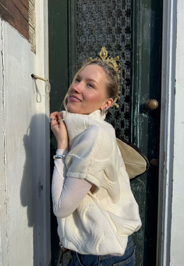 Joven mujer con un suéter de punto blanco de pie frente a una puerta oscura y ornamentada, sonriendo y mirando hacia arriba, con la luz del sol proyectando una sombra en la pared.