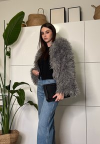 Jeune femme portant une veste grise duveteuse par-dessus un haut noir et un jean bleu, tenant une pochette noire, debout à côté de grandes plantes d'intérieur vertes.