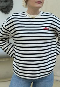 Ριγέ πουλόβερ με μαύρες και κρεμ οριζόντιες γραμμές, με το "Merci" κεντημένο σε κόκκινο. Συνδυάζεται με ανοιχτό μπλε τζιν. Κανονική εφαρμογή.