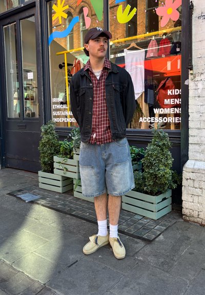 Persona vestida con una chaqueta de denim negra sobre una camisa de cuadros roja, pantalones cortos de denim azul claro, calcetines blancos y zapatos beige, de pie frente a una tienda.