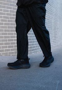 Personne portant un pantalon de sport noir et des baskets noires marchant sur un trottoir avec un mur en briques grises en arrière-plan.