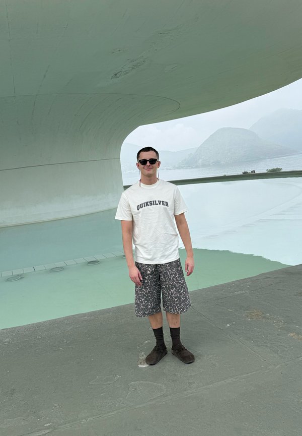 Junger Mann mit Sonnenbrille, weißem Quiksilver-Shirt, gemusterten Shorts und dunklen Schuhen steht nahe einer modernen, geschwungenen Struktur am Wasser mit entfernten Hügeln im Hintergrund.