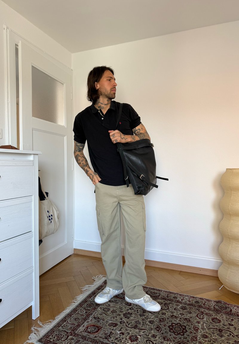 Homme avec des tatouages portant un polo noir, un pantalon beige, des baskets blanches, portant un sac à dos en cuir noir dans une pièce lumineuse avec un sol en bois et un tapis à motifs.