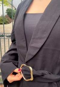 Jachetă neagră tip peliculă, realizată dintr-un material texturat, cu reveruri late și o cataramă rectangulară unică aurie în talie.