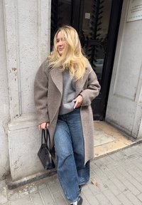 Cappotto lungo in lana marrone sopra un maglione grigio, abbinato a jeans larghi blu e scarpe nere. Tiene una piccola borsa nera.