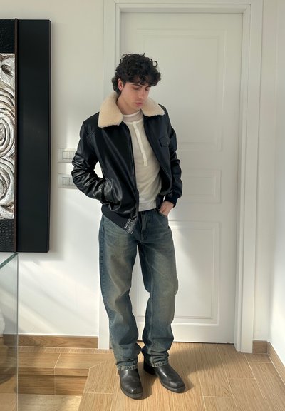 Chaqueta bomber de cuero negro con cuello de piel crema, camiseta blanca tipo henley y jeans anchos azules, combinados con botas negras de tobillo. Fondo neutro.