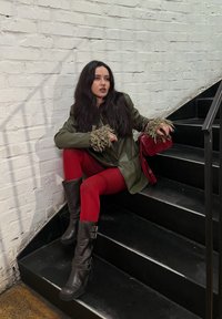 Blazer verde oliva con puños texturizados y esponjosos, leggings rojos y botas de cuero marrón hasta la rodilla. Pequeño bolso rojo. Fondo de escalera negra.