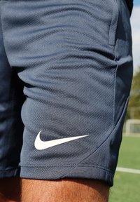 Detailní záběr na nohu sportovce oblečenou v tmavě modrých strukturovaných sportovních šortkách Nike na zeleném hřišti s brankou v rozmazaném pozadí.