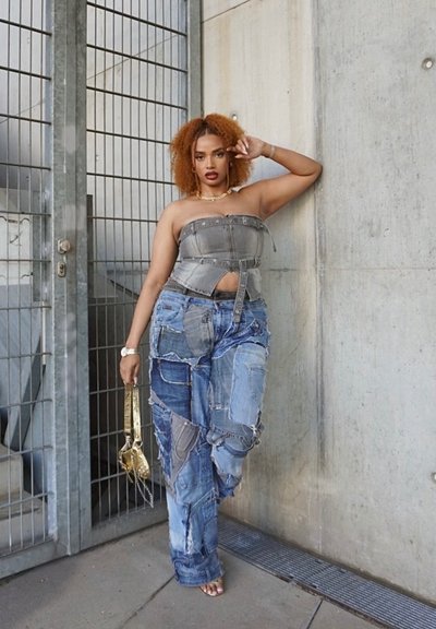 Top bandeau de mezclilla gris combinado con pantalones anchos de mezclilla azul con parcheo. Sostiene un bolso de cadena dorada. La textura está desgastada, acentuando el diseño en capas.