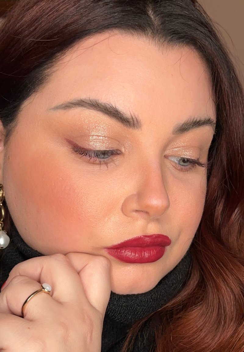 Portrait d'une femme aux cheveux ondulés rouge foncé, portant un col roulé noir, des boucles d'oreilles en or avec des perles et un rouge à lèvres rouge vif.