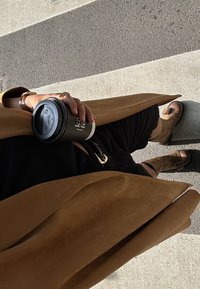 Cappotto in suede tan drappeggiato, pantaloni corti neri, scarpe basse marroni con fibbie, mentre tiene una tazza di caffè nero con un'etichetta di avviso. Pavimentazione a strisce sotto.