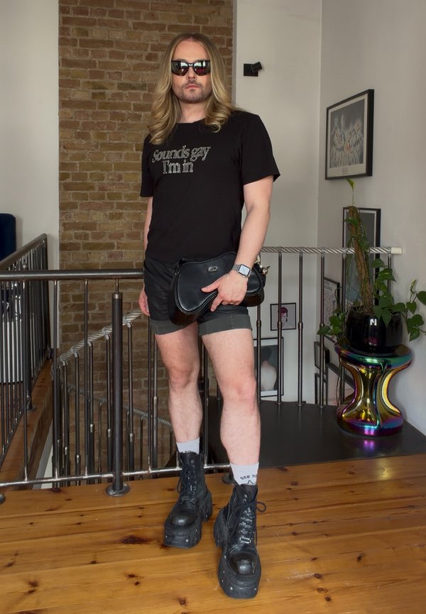 Personne aux longs cheveux blonds portant des lunettes de soleil, un t-shirt noir avec texte, un short noir, des bottes à plateforme noires, tenant un sac à main noir à l'intérieur.
