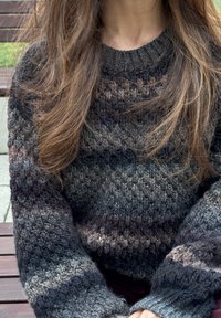 Νεαρό άτομο με μακριά ανοικτή καστανή τρίχα που φοράει χοντρό πλεκτό πουλόβερ με οριζόντιες ρίγες σε σκούρο γκρι και καφέ, καθισμένο σε ξύλινο πάγκο.