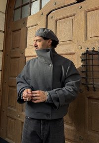 Homme portant une veste grise et un béret, debout les mains jointes, regardant vers la gauche devant une grande porte en bois avec des accents métalliques.