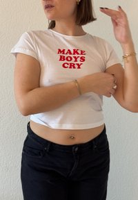 Bela skrajšana majica z rdečim napisom "MAKE BOYS CRY", v kombinaciji s črnimi hlačami visoko pasu in srebrnim zapestnikom na zapestju.