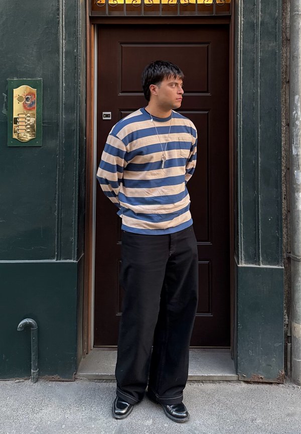 Striped long-sleeve shirt in beige and blue, black wide-leg pants, black shiny shoes, standing beside a dark wooden door with a brass intercom.