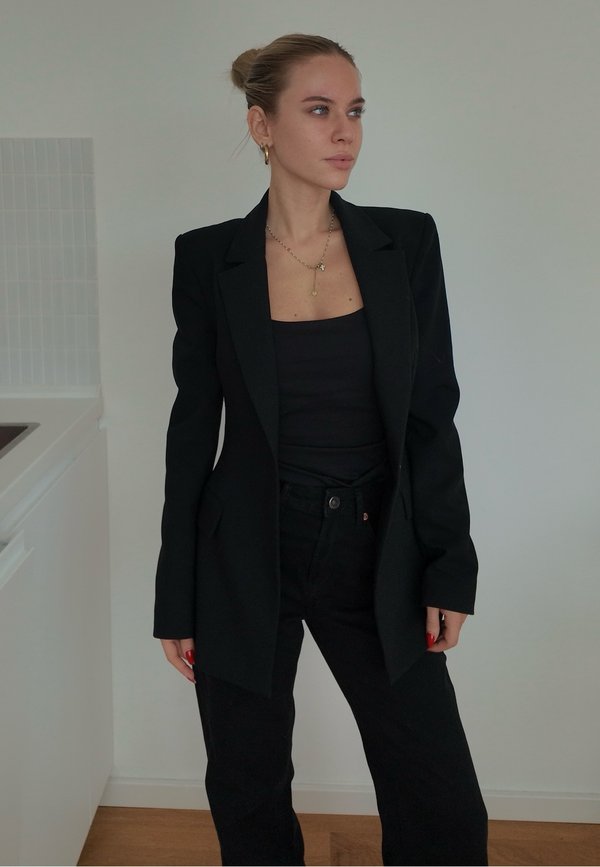 Une femme pose à l’intérieur, vêtue d’un blazer noir, d’un haut et d’un pantalon, avec des bijoux en or et les cheveux relevés en chignon.