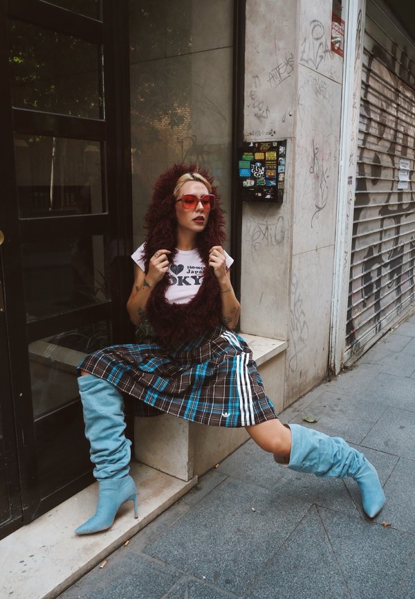 Femme portant des lunettes de soleil teintées rouges, une capuche en fourrure, une jupe à carreaux et des bottes bleues à talons hauts, assise avec les jambes étirées sur un trottoir urbain.