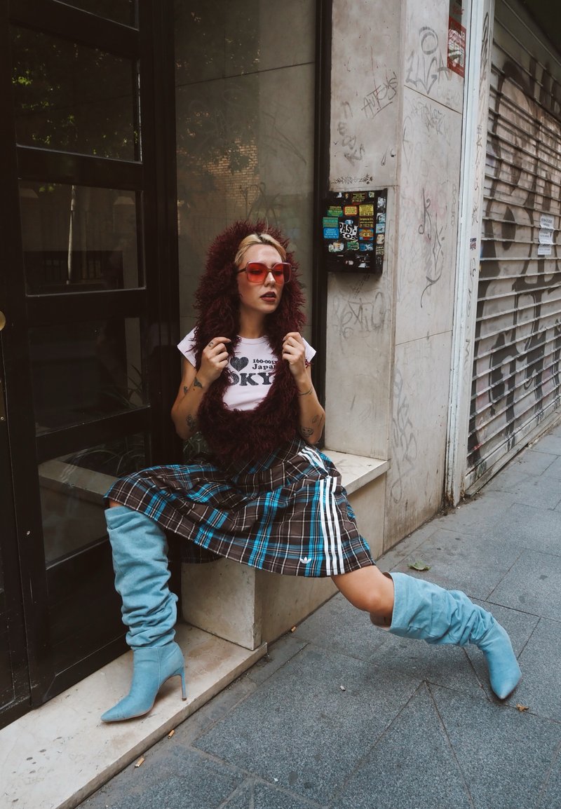 Femme portant des lunettes de soleil teintées rouges, une capuche en fourrure, une jupe à carreaux et des bottes bleues à talons hauts, assise avec les jambes étirées sur un trottoir urbain.