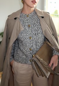 Manteau trench beige sur un pull en maille gris avec des boutons dorés, associé à un pantalon beige clair et un sac en cuir marron avec des accents cousus.