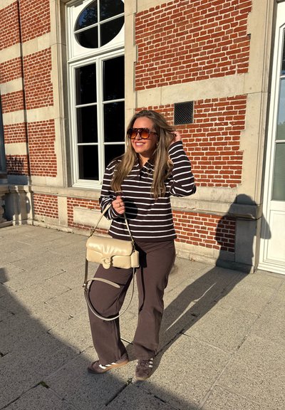 Top de manga larga a rayas en marrón oscuro y blanco, combinado con pantalones marrones y zapatillas grises. Sosteniendo un bolso de hombro beige con un diseño curvado.