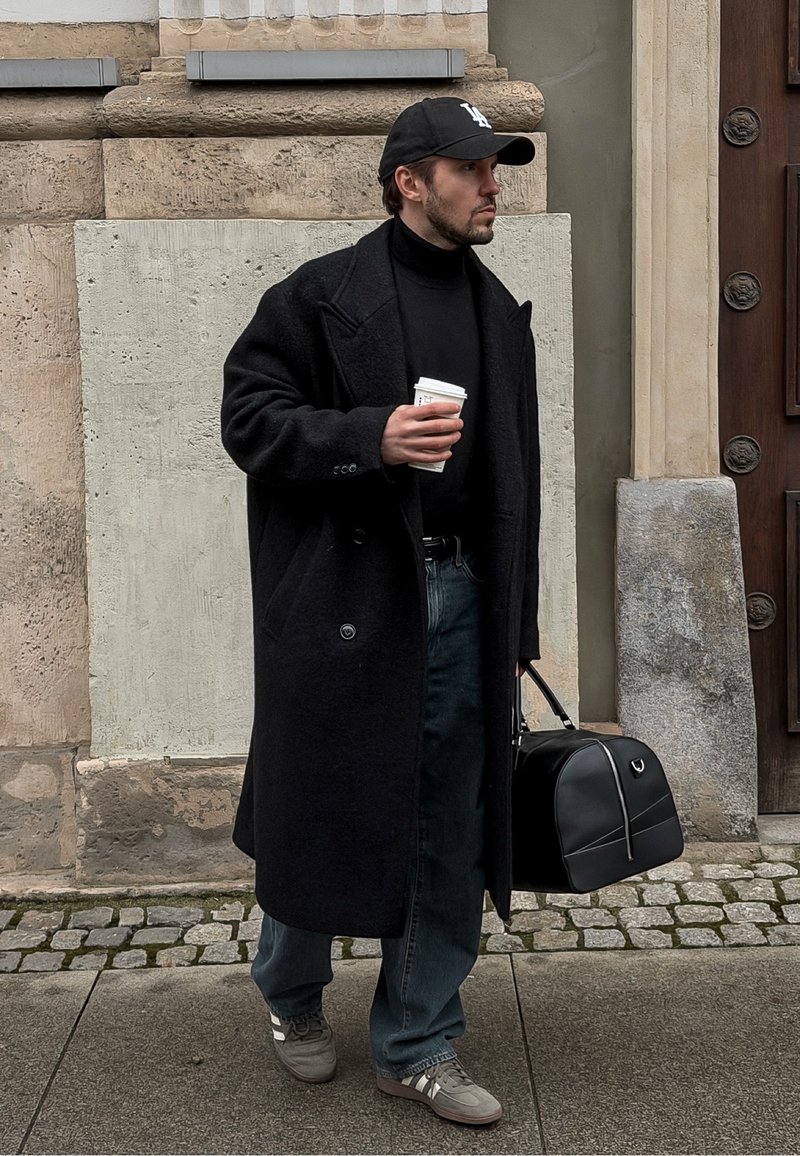 Manteau en laine noire avec de grands revers, col roulé noir, jean en denim foncé et baskets grises. Tenant une tasse de café et un sac noir.