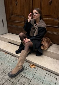 Μαύρο παλτό, καφέ πλεκτό περιτύλιγμα, μαύρη φόρεμα, διαφανείς μαύρες καλσόν, ταμπά μπότες μέχρι το γόνατο με καουτσούκ σόλες, καθισμένες σε πέτρινες σκάλες με πλακάκια από κάτω.