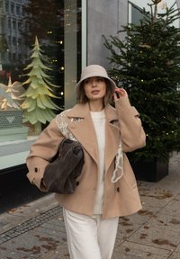 Béžový vlnený kabát s dekoratívnou bielou výšivkou, zladený so svetlým svetrom a tmavou kabelkou, doplnený sivým plsteným klobúkom.