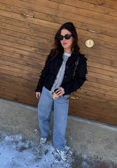 Chaqueta negra texturizada, camisa gris con botones, vaqueros azul claro y zapatillas deportivas. Sosteniendo una taza de café. Fondo de madera y nieve.