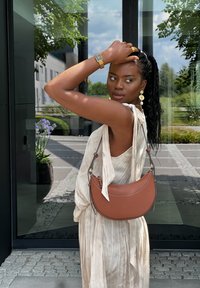 Sac à bandoulière en cuir marron au design courbé, avec sangle réglable et couture discrète. Modèle portant une robe légère et fluide avec des plis.