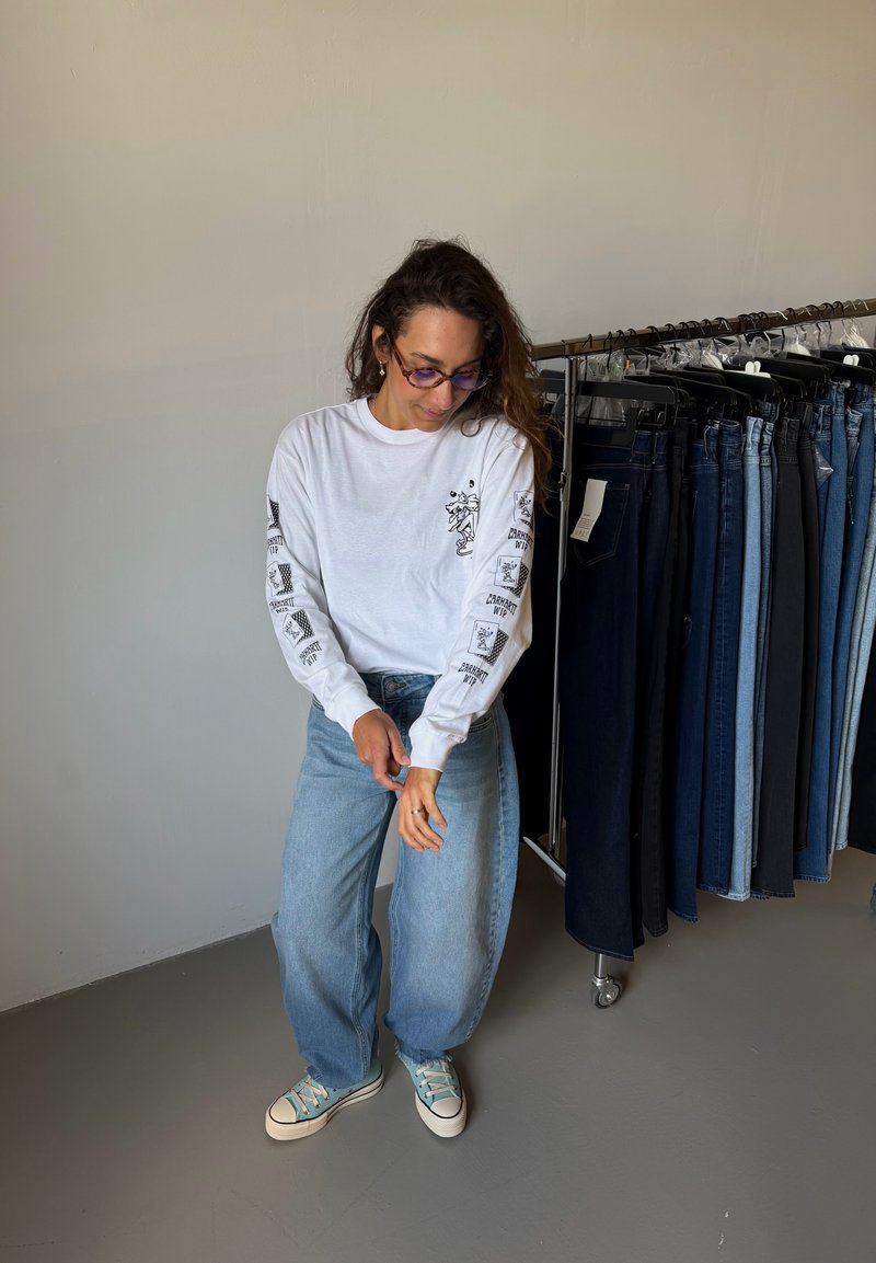 Femme portant une chemise blanche à manches longues et un jean bleu ajuste la manche près d'un portant avec différents pantalons en denim sur des cintres.