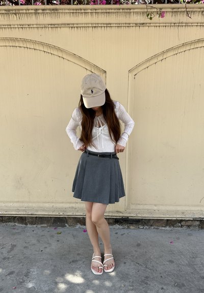 Falda gris plisada con cinturón negro, combinada con una blusa de manga larga blanca con un detalle de lazo. Una gorra beige y sandalias blancas completan el conjunto.