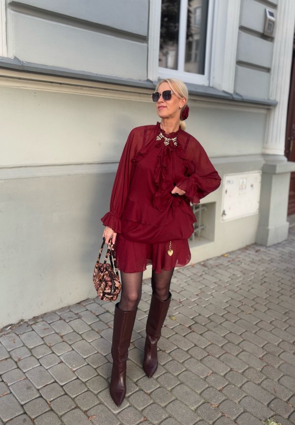 Una mujer con un vestido burdeos con volantes, gafas de sol oscuras y botas altas posa en una calle empedrada con un bolso estampado.
