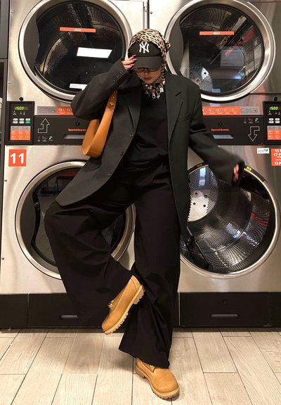 Blazer negro de gran tamaño, top negro, pantalones anchos negros. Zapatos marrón claro, bolso cruzado marrón. Máquinas de lavandería al fondo con pantallas digitales.