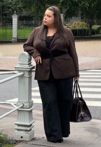 Bruine blazer met een strikriem over een zwarte top, gecombineerd met donkere jeans en zwarte schoenen. Houdt een donkere handtas vast, leunend tegen een leuning.
