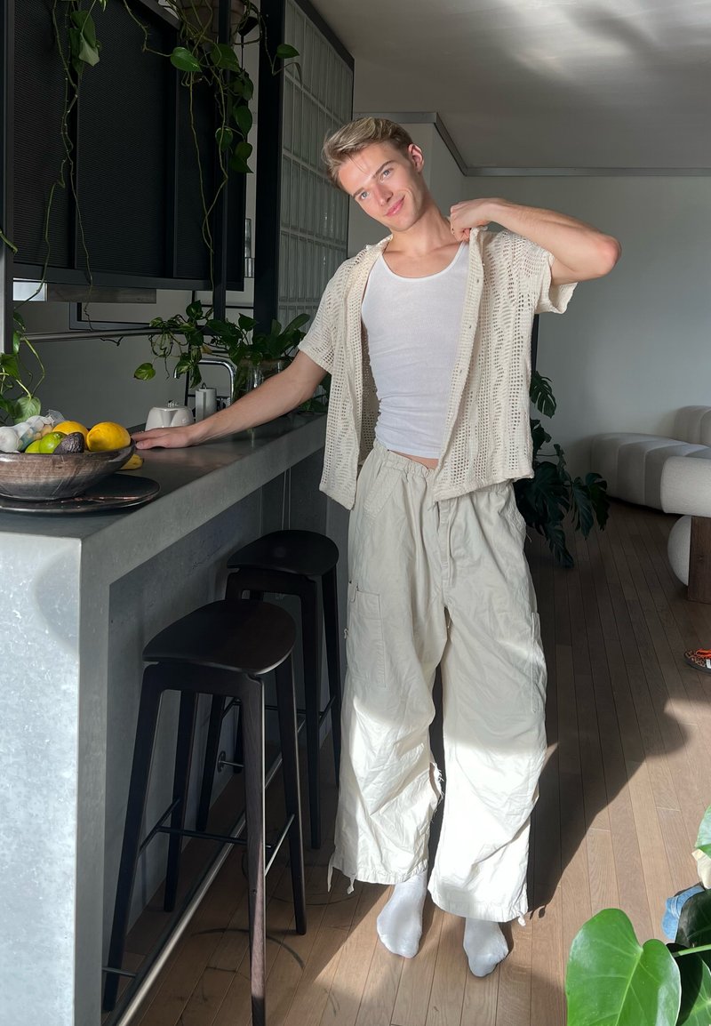 Giovane persona in piedi scalza in una cucina moderna illuminata dal sole, indossa pantaloni bianchi larghi, una canottiera e una camicia aperta a maniche corte.