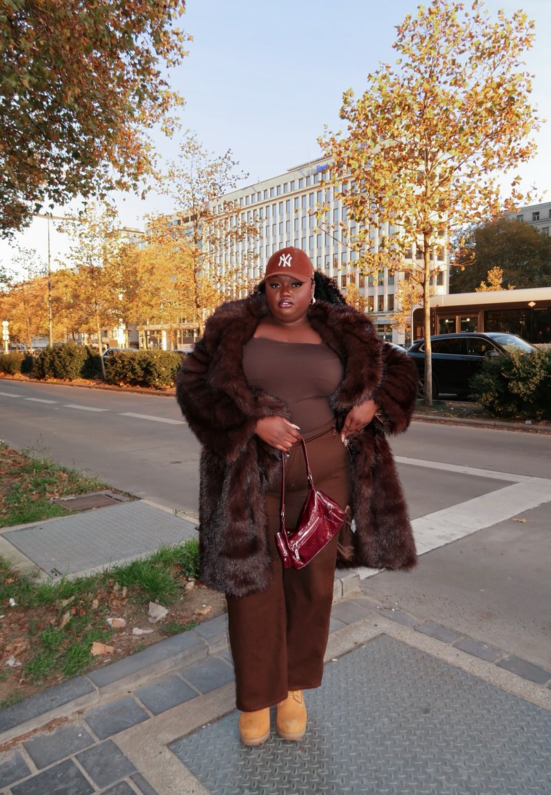 Manteau en fourrure marron, haut marron ajusté et pantalon assorti. Sac à main bordeaux, bottes marron et casquette marron. Décor urbain avec des arbres et des bâtiments.