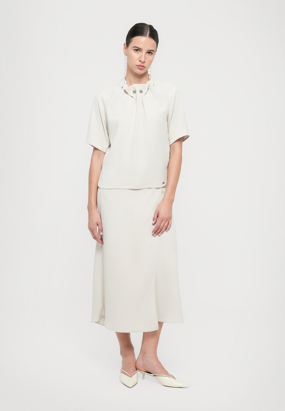 Blouse beige clair à manches courtes avec un col à volants et trois boutons décoratifs, associée à une jupe longueur cheville. Coupe ajustée, texture lisse.