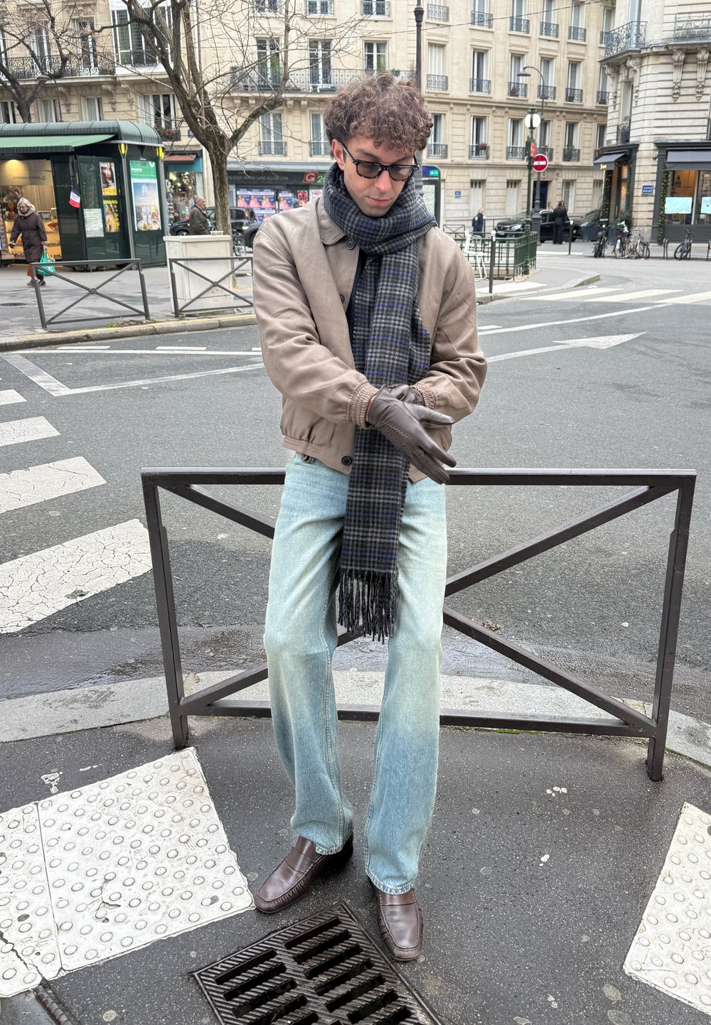 Άνδρας με γυαλιά, καφέ μπουφάν, κασκόλ και γάντια στέκεται σε γωνία δρόμου της πόλης κοντά σε διάβαση πεζών και μεταλλική κιγκλίδα.