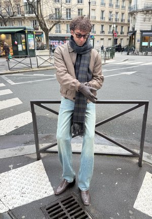 Hombre con gafas, chaqueta marrón, bufanda y guantes, de pie en la esquina de una calle de la ciudad cerca de un paso de peatones y una barandilla metálica.