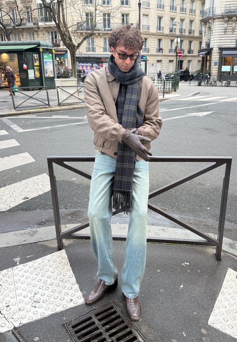 Homme portant des lunettes, veste marron, écharpe et gants, debout au coin d'une rue en ville près d'un passage piéton et d'une barrière métallique.