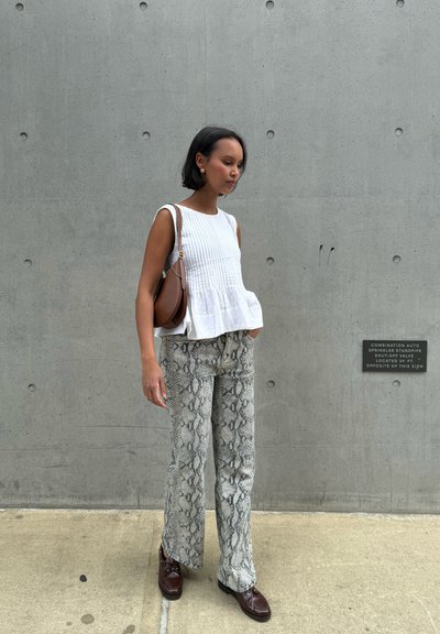 Top sin mangas blanco y texturizado con dobladillo de peplum; pantalones de pierna ancha gris con estampado de serpiente; bolso de hombro de cuero marrón; zapatos marrón oscuro. Fondo de hormigón.
