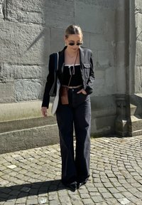 Mustahdhi blazer mweusi wenye vifungo, koti la juu la mweusi lililo na kamba, jeans za juu za mweusi, na begi nyepesi, dhidi ya ukuta wa jiwe.