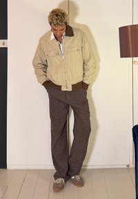 Νεαρό άτομο με κατσαρά μαλλιά, που φοράει μπεζ σακάκι με λεπτές ρίγες, καφέ παντελόνια και γκρι αθλητικά παπούτσια, στέκεται μέσα σε εσωτερικό χώρο με τα χέρια στις τσέπες.