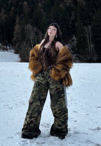 Jeune femme debout dans un champ enneigé, portant un manteau en fourrure, un haut imprimé léopard, un pantalon camouflage et des chaussures foncées, avec une forêt en arrière-plan.