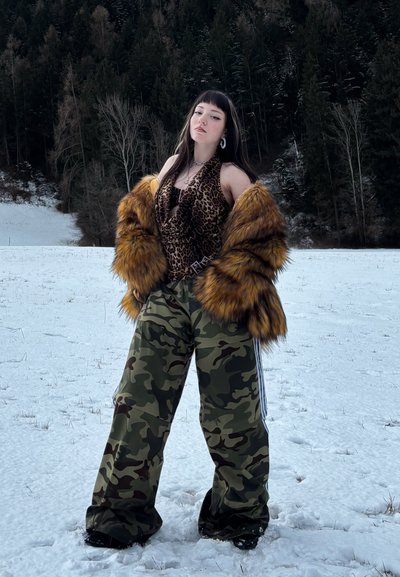 Joven de pie en un campo nevado, vestida con abrigo de piel, blusa con estampado de leopardo, pantalones de camuflaje y zapatos oscuros, con un fondo de bosque.