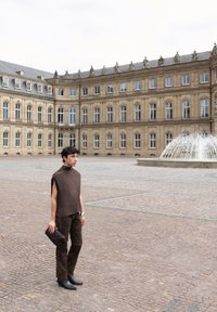 Persoon staand op een kasseienplein bij een fontein, gekleed in een bruin mouwloos topje en broek, met een donkere gevlochten clutch in de hand.